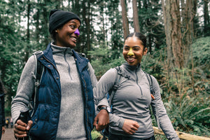 Two women arm-in-arm wearing colorful Nöz sunscreen on their noses while walking through the woods. 